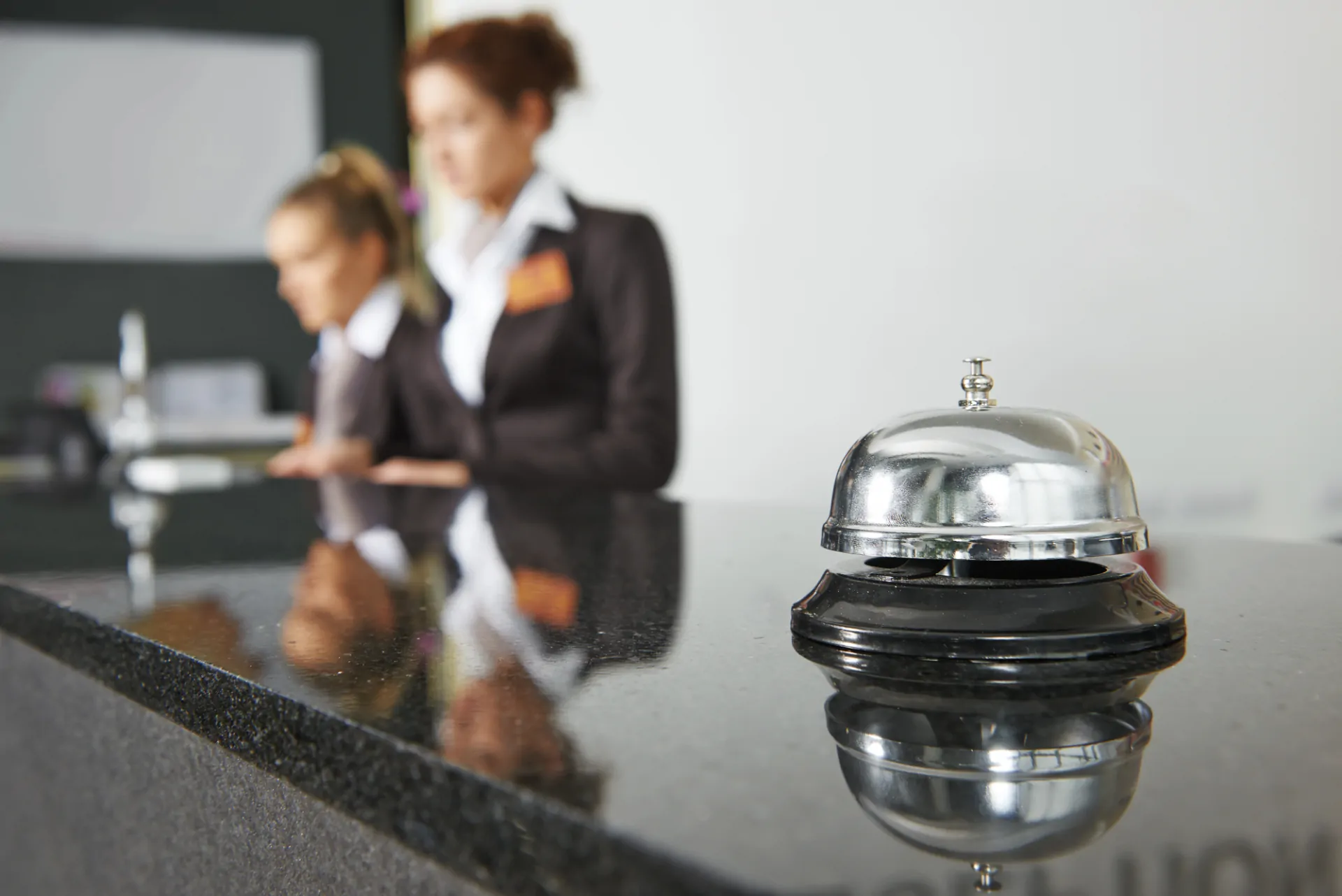 Visuale ravvicinata di un bancone di hotel con primo piano di campanello e receptionist sullo sfondo intente a informarsi sulla sicurezza sul lavoro in hotel